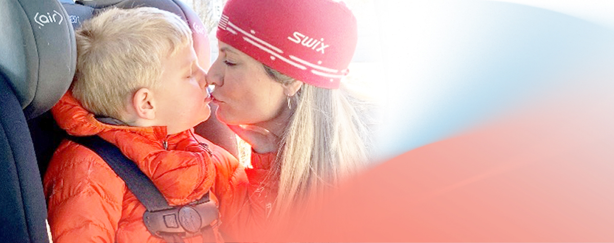 Child in orange jacket sits in a car seat beside someone wearing a red Swix hat.